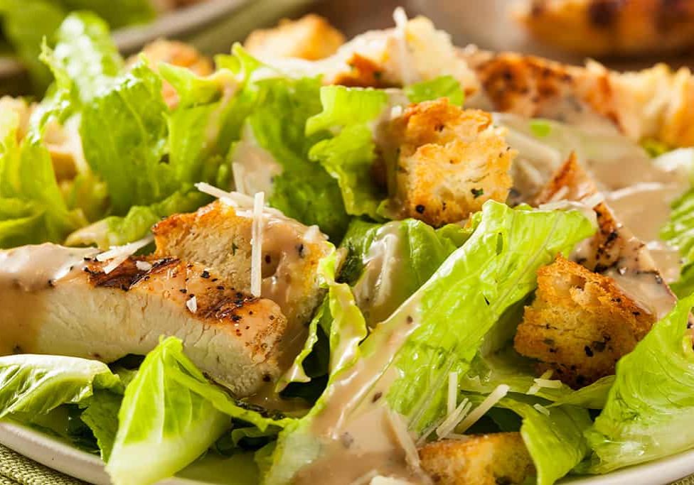 A close-up of a caesar salad with grilled chicken, croutons, and shredded parmesan, drizzled with creamy dressing.